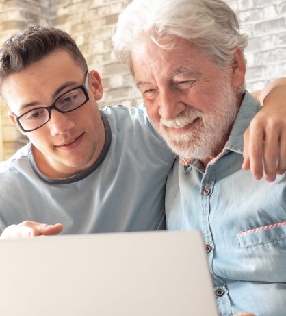 Smiling experienced young boy with laptop teaching and showing n Smiling experienced young boy with laptop teaching and showing new computer technology to his old grandfather for surfing internet, shopping online, managing bank. Old man learns to use the computer