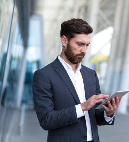 stylish bearded businessman in formal business suit standing working with tablet in hands on background modern office building outside. Man using smartphone or uses mobile phone outdoors city street stylish bearded businessman in formal business suit standing working with tablet in hands on background modern office building outside. Man using smartphone or uses mobile phone outdoors city street